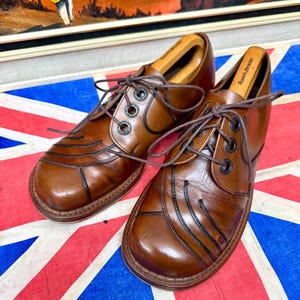 May include: A pair of brown leather oxford shoes featuring dark brown stitched detailing on the toe box, black eyelets, and thin brown laces. Each shoe contains a wooden shoe tree with the brand name Russell & Bromley printed on the heel.