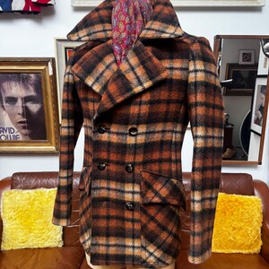 May include: A vintage double-breasted plaid coat in shades of brown, orange, and black. The coat features a wide collar, two rows of buttons, and two front pockets. A paisley scarf is draped over the collar. The coat is displayed on a mannequin.