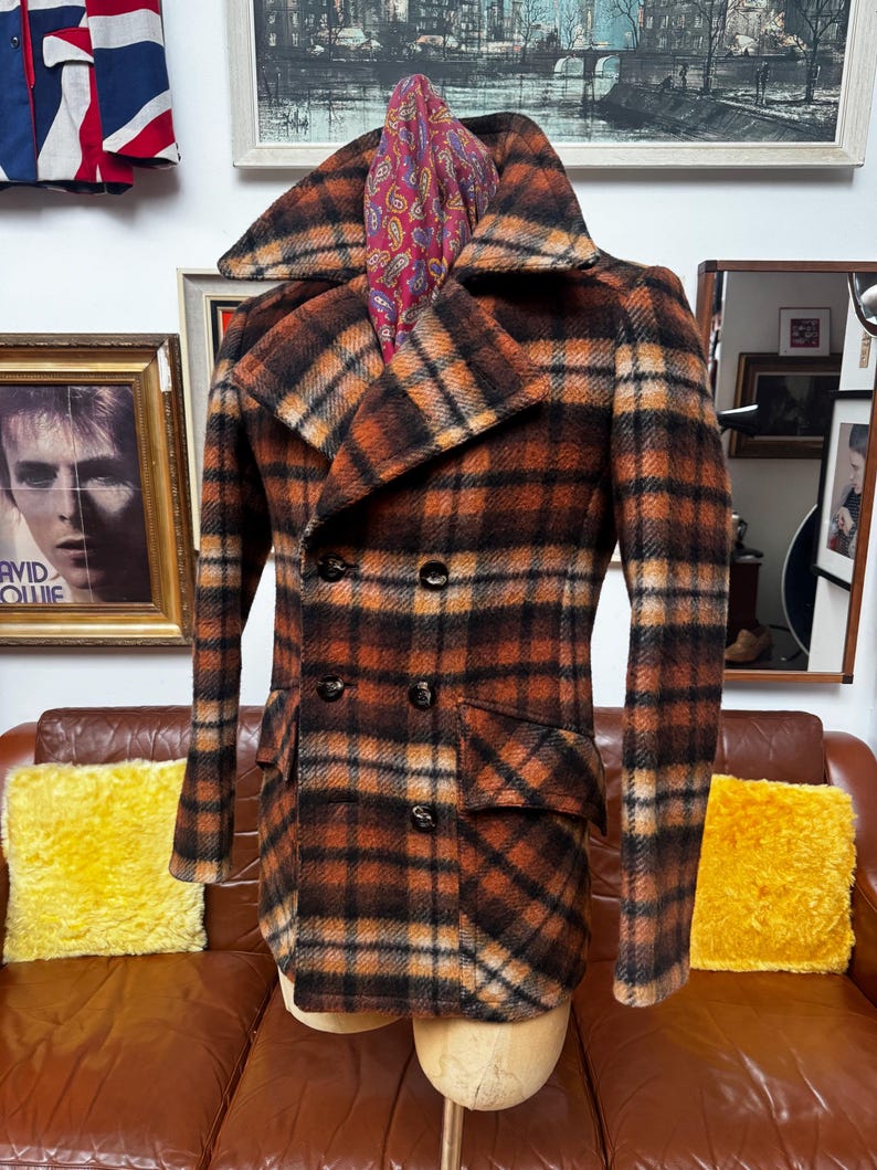 May include: A vintage double-breasted plaid coat in shades of brown, orange, and black. The coat features a wide collar, two rows of buttons, and two front pockets. A paisley scarf is draped over the collar. The coat is displayed on a mannequin.