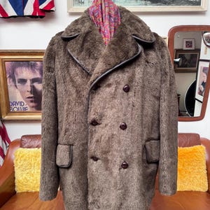 May include: A brown faux fur coat with a double-breasted design. The coat features dark brown buttons, a notched collar, and dark brown trim around the pockets and lapels. A patterned scarf is draped over the coat.