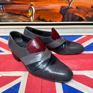 K&ouml;nnte beinhalten: Ein Paar Vintage-Schuhe aus grauem Leder mit geometrischem Design. Die Schuhe haben einen bordeauxfarbenen Akzent auf der Oberseite und ein schwarz-graues Muster auf dem Blatt. Die Schuhe haben einen kleinen Absatz und eine runde Spitze.