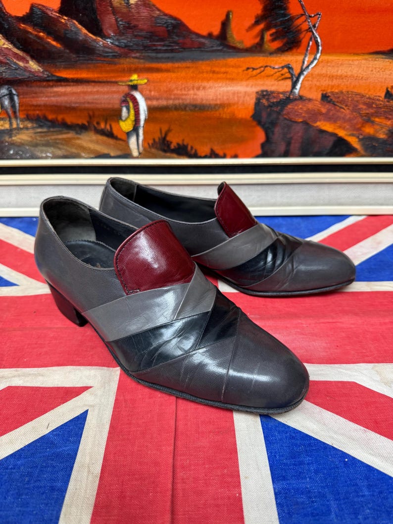 K&ouml;nnte beinhalten: Ein Paar Vintage-Schuhe aus grauem Leder mit geometrischem Design. Die Schuhe haben einen bordeauxfarbenen Akzent auf der Oberseite und ein schwarz-graues Muster auf dem Blatt. Die Schuhe haben einen kleinen Absatz und eine runde Spitze.