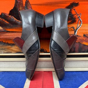 K&ouml;nnte beinhalten: Ein Paar graue und schwarze Lederschuhe mit Blockabsatz. Die Schuhe haben ein Colorblock-Design mit einem burgunderfarbenen Akzent. Die Schuhe werden auf einer Union Jack-Flagge pr&auml;sentiert.