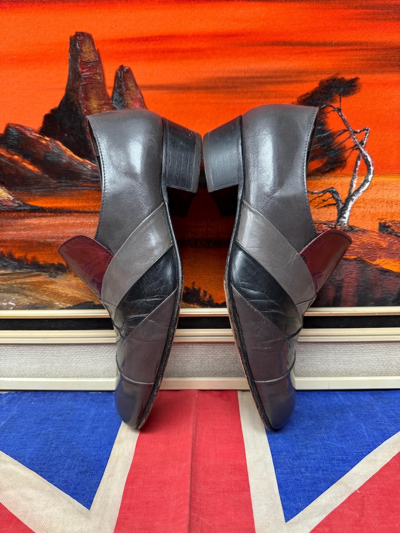 K&ouml;nnte beinhalten: Ein Paar graue und schwarze Lederschuhe mit Blockabsatz. Die Schuhe haben ein Colorblock-Design mit einem burgunderfarbenen Akzent. Die Schuhe werden auf einer Union Jack-Flagge pr&auml;sentiert.