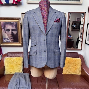 May include: A blue plaid suit jacket and matching trousers displayed on a mannequin. The jacket has a burgundy paisley pocket square and scarf. The suit is displayed on a brown leather couch with yellow pillows.
