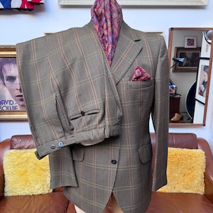May include: A brown plaid suit with a matching jacket and pants, displayed on a mannequin. The jacket has a pocket square and a paisley scarf is draped around the neck. The suit is a classic style.