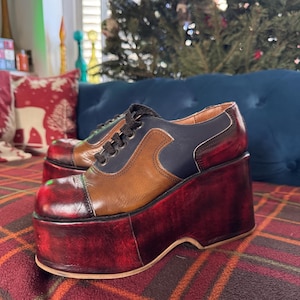 May include: Platform shoes with a multi-tone design. The shoes feature a brown, navy blue, and red color scheme. The platform soles are a deep red, and the uppers combine brown and navy blue leather. Black laces complete the look.