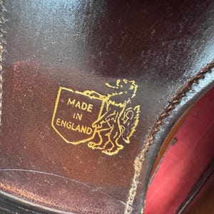 May include: Close-up of a brown leather shoe sole. A gold emblem with the words "MADE IN ENGLAND" and a heraldic lion is visible. The shoe's stitching and the red interior are also partially visible.