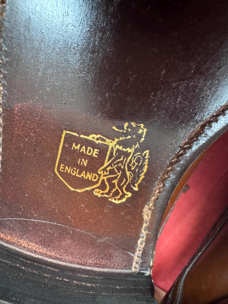 May include: Close-up of a brown leather shoe sole. A gold emblem with the words "MADE IN ENGLAND" and a heraldic lion is visible. The shoe's stitching and the red interior are also partially visible.