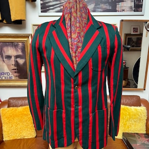 Puede incluir: Una blazer verde con llamativas rayas verticales rojas. La chaqueta tiene un diseño clásico con solapa de muesca, cierre de un botón y dos bolsillos delanteros. Una bufanda estampada está drapeada sobre el maniquí. El blazer se exhibe en un maniquí.