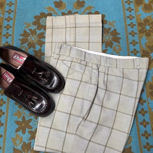 May include: A pair of brown leather tassel loafers and light gray plaid trousers are displayed on a patterned blue and brown rug. The trousers are folded, and the shoes are positioned near the pants.