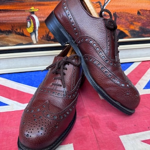 May include: A pair of dark brown leather brogue shoes featuring a textured pebble grain finish, decorative perforated detailing along the seams, a rounded toe, and dark brown laces. The shoes have a stacked heel and a black sole.