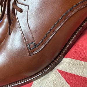 May include: Close-up of a pair of brown leather dress shoes with dark brown laces. The shoes have decorative stitching and a classic design. The shoes are on a patterned fabric with red, white, and blue colors.