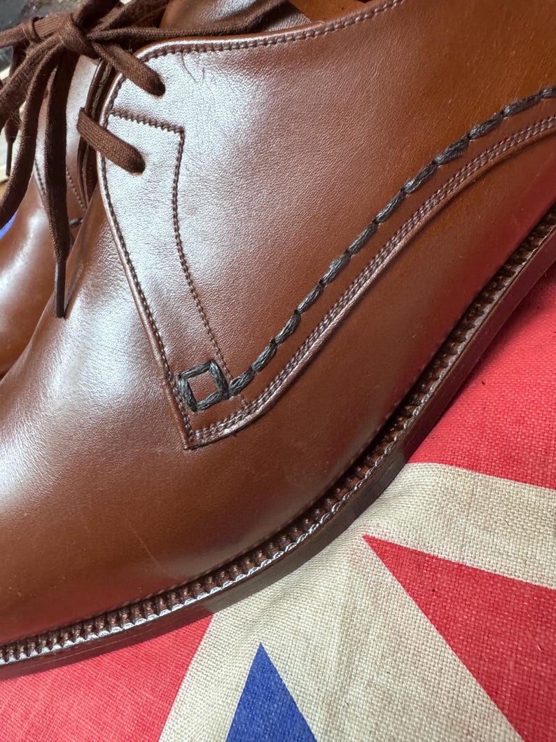 May include: Close-up of a pair of brown leather dress shoes with dark brown laces. The shoes have decorative stitching and a classic design. The shoes are on a patterned fabric with red, white, and blue colors.