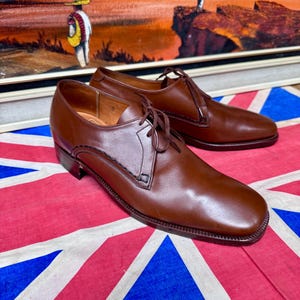 May include: A pair of brown leather dress shoes with laces. The shoes are displayed on a Union Jack flag. The shoes have a classic design with a square toe and a low heel. The background features a framed painting.