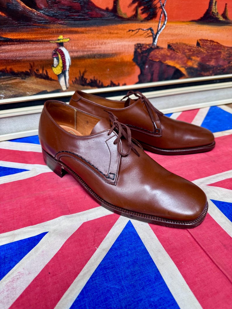 May include: A pair of brown leather dress shoes with laces. The shoes are displayed on a Union Jack flag. The shoes have a classic design with a square toe and a low heel. The background features a framed painting.
