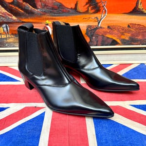 May include: A pair of black leather Chelsea boots featuring a pointed toe, a stacked heel, and black elastic side panels. The boots have a smooth, polished finish and a pull tab at the back of the ankle collar for ease of wear and a sleek silhouette.