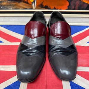 K&ouml;nnte beinhalten: Ein Paar graue Lederschuhe mit geometrischem Design. Die Schuhe haben eine weinrote Kappe, graue und schwarze Chevron-Akzente und eine abgerundete Spitze. Die Schuhe werden auf einer Union Jack-Flagge pr&auml;sentiert.