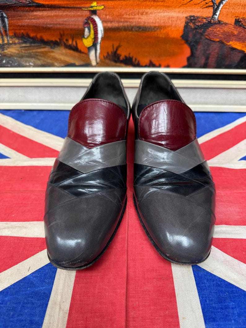 K&ouml;nnte beinhalten: Ein Paar graue Lederschuhe mit geometrischem Design. Die Schuhe haben eine weinrote Kappe, graue und schwarze Chevron-Akzente und eine abgerundete Spitze. Die Schuhe werden auf einer Union Jack-Flagge pr&auml;sentiert.