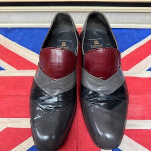 K&ouml;nnte beinhalten: Ein Paar Herren-Anzugschuhe mit einem einzigartigen Design. Die Schuhe weisen ein Farbblockmuster mit Abschnitten aus bordeauxfarbenem, grauem, schwarzem und dunkelgrauem Leder auf. Die Schuhe werden auf einer Union Jack-Flagge pr&auml;sentiert.