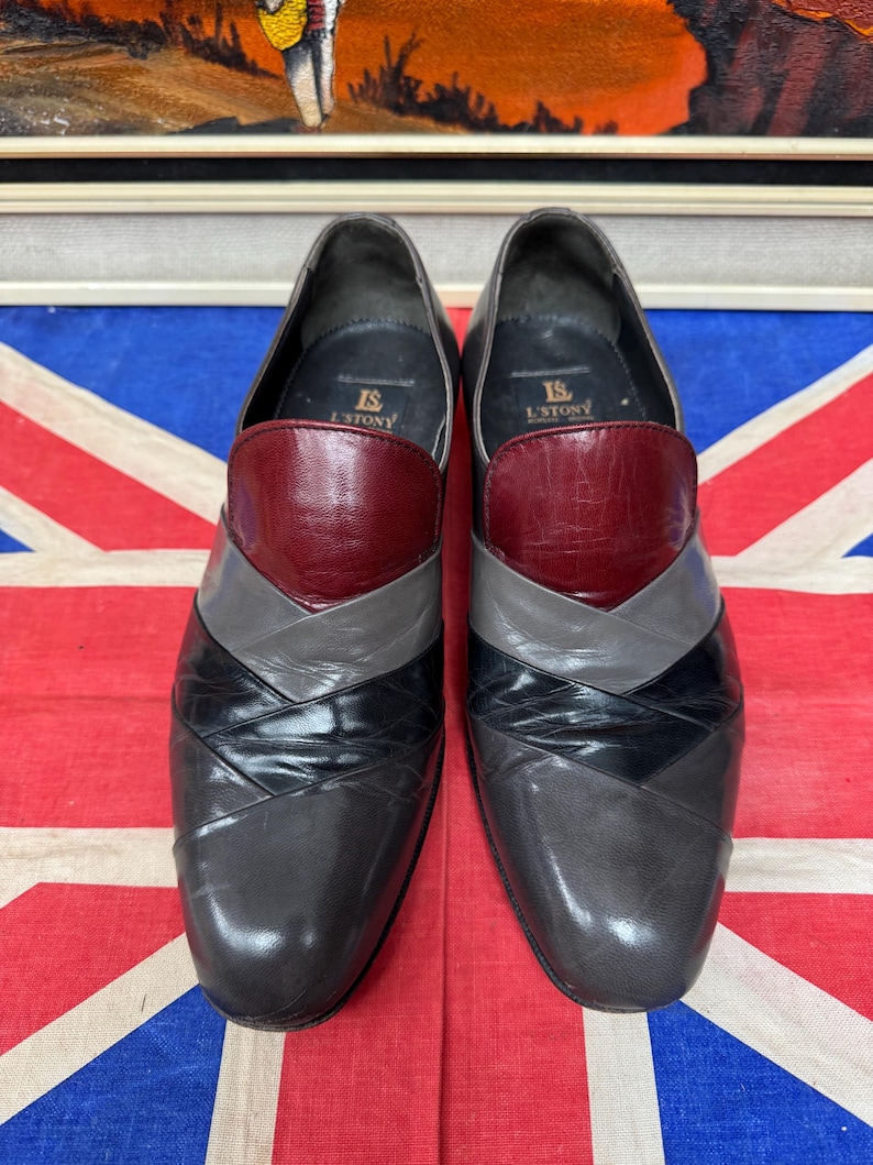 K&ouml;nnte beinhalten: Ein Paar Herren-Anzugschuhe mit einem einzigartigen Design. Die Schuhe weisen ein Farbblockmuster mit Abschnitten aus bordeauxfarbenem, grauem, schwarzem und dunkelgrauem Leder auf. Die Schuhe werden auf einer Union Jack-Flagge pr&auml;sentiert.