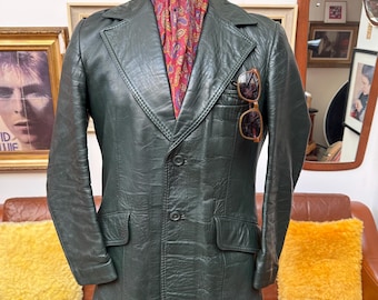 Pode incluir: Um blazer de couro verde-escuro com lapela entalhada, dois botões frontais e dois bolsos com aba. Um lenço com padrão paisley vermelho e dourado está inserido no decote e óculos de sol com armação castanha estão presos no bolso do peito.