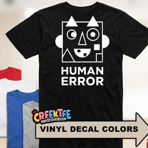 May include: Black t-shirt with a white graphic of a robot face and the words "HUMAN ERROR". The image also shows a variety of vinyl decal colors and folded t-shirts in red, white, blue, and gray.