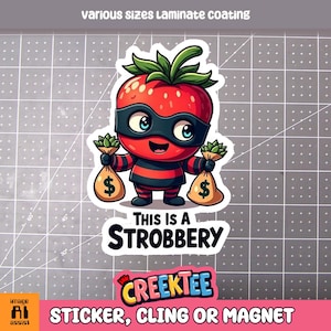 May include: A red cartoon strawberry wearing a black mask and holding two bags of money. The text "This is a Strobbery" is written below the strawberry. The text "CREEKTEE" is written below the text. The text "STICKER, CLING OR MAGNET" is written below the text.