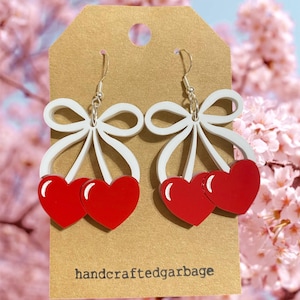 May include: A pair of dangle earrings featuring a red heart design with a white bow accent. The earrings are attached to silver-colored hooks. The earrings are displayed on a brown tag with the text "handcraftedgarbage".