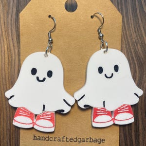 May include: A pair of white ghost-shaped earrings with black eyes and a curved mouth, wearing red sneakers. The earrings are hanging from silver hooks. The text "handcraftedgarbage" is printed on the brown tag.