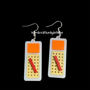 May include: A pair of earrings with a white, yellow, orange, and red design. The earrings are shaped like a rectangle with a small hole at the top for the earring hook. The earrings are made of acrylic and have a textured surface.
