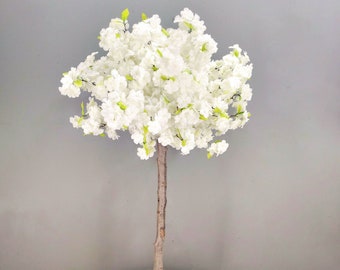Könnte beinhalten: Ein künstlicher Kirschblütenbaum in Weiß mit braunem Stamm und grünen Blättern. Der Baum steht auf einem weißen Marmortisch mit schwarzen Beinen. Eine kleine grüne Pflanze befindet sich in einem geflochtenen Korb neben dem Tisch. Ein Schild mit dem Wort "Wald" lehnt an dem Korb.