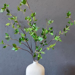 May include: A white ceramic vase holds a collection of faux greenery. The branches are brown with small, bright green leaves. The vase has a rounded shape and a narrow neck, set against a gray background.