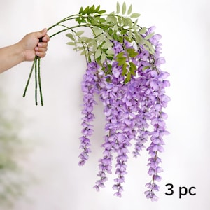 May include: Artificial wisteria stems with cascading purple flowers and green leaves. The stems are held in a hand, with the text "3 pc" in the lower right corner. Ideal for home decor or floral arrangements.