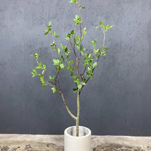 May include: A small artificial tree with green leaves and brown branches in a white cylindrical pot. The tree is standing on a gray concrete floor.