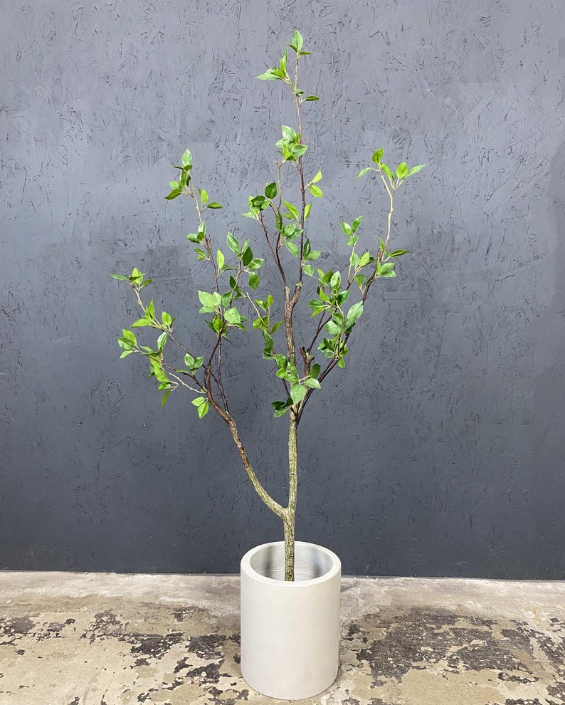 May include: A small artificial tree with green leaves and brown branches in a white cylindrical pot. The tree is standing on a gray concrete floor.