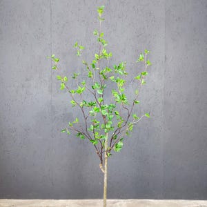 May include: A green artificial tree with a brown trunk in a black pot. The tree is in front of a gray wall.
