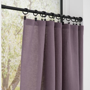 May include: A set of purple linen curtains hanging from a black rod with rings. The curtains are partially open, revealing a blurred view of a tree and foliage outside the window. The curtains are a solid color and appear to be made of a lightweight fabric.