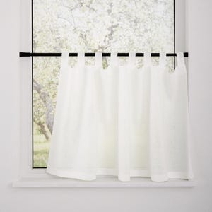May include: White linen curtain hanging on a black rod inside a white window frame. The curtain is semi-sheer, allowing light to filter through. The window looks out onto a blurred view of a tree with green leaves.