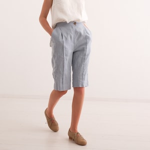 May include: A woman wearing a white sleeveless top and light blue linen shorts. The shorts have a button closure and pockets. She is wearing brown leather shoes. The outfit is simple and elegant, suitable for warm weather.