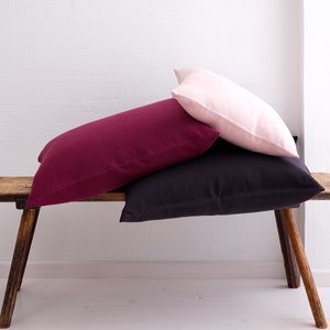 A pile of deocorative square pillow covers, in the colors pale pink, wine red and dark violet with a hidden zipper closure on a wooden bench.