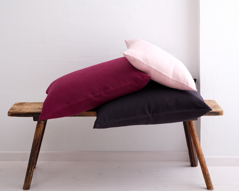 A pile of deocorative square pillow covers, in the colors pale pink, wine red and dark violet with a hidden zipper closure on a wooden bench.