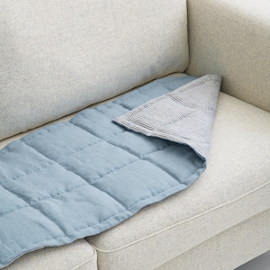 K&ouml;nnte beinhalten: Eine blau gesteppte Decke mit einem gestreiften Stoff, der dar&uuml;ber gefaltet ist, liegt auf einem hellen Sofa. Die Decke hat eine rechteckige Form mit quadratischen N&auml;hten, und der gefaltete Stoff hat ein blau-wei&szlig;es Streifenmuster.
