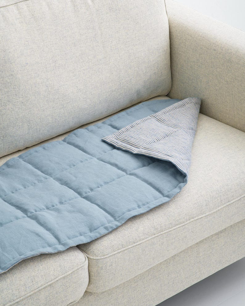 K&ouml;nnte beinhalten: Eine blau gesteppte Decke mit einem gestreiften Stoff, der dar&uuml;ber gefaltet ist, liegt auf einem hellen Sofa. Die Decke hat eine rechteckige Form mit quadratischen N&auml;hten, und der gefaltete Stoff hat ein blau-wei&szlig;es Streifenmuster.