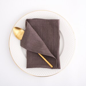 May include: A brown linen napkin folded into a triangular shape on a white plate with a gold rim. A gold spoon is placed on top of the napkin.