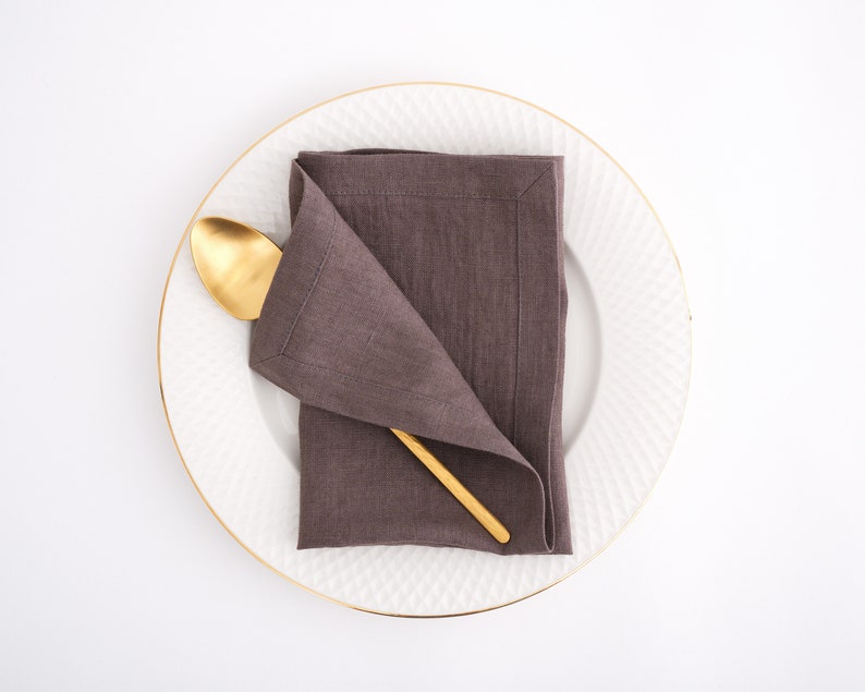 May include: A brown linen napkin folded into a triangular shape on a white plate with a gold rim. A gold spoon is placed on top of the napkin.