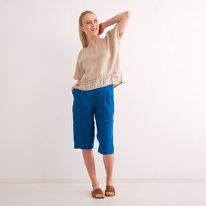 May include: A woman wearing a beige linen top and bright blue linen capri pants. The top has short sleeves and a layered design. The capri pants are knee-length with vertical seams. The woman is wearing brown sandals.