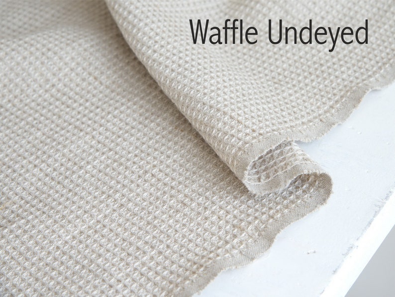 Pode incluir: Close-up de um tecido de waffle em uma cor natural n&atilde;o tingida. O tecido est&aacute; dobrado sobre si mesmo, mostrando a textura e a trama. O texto "Waffle Undyed" &eacute; vis&iacute;vel no canto superior direito da imagem.
