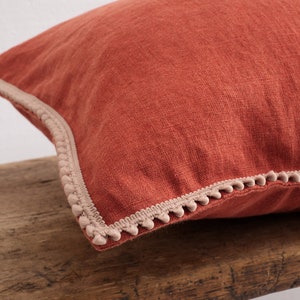 May include: A rust-colored linen pillow with a beige pom-pom trim.