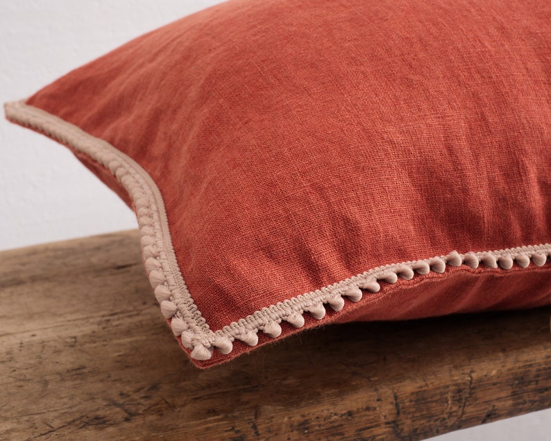 May include: A rust-colored linen pillow with a beige pom-pom trim.